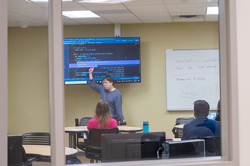  A professor gesturing at a computer monitor while teaching a class, observed from outside through a glass window.