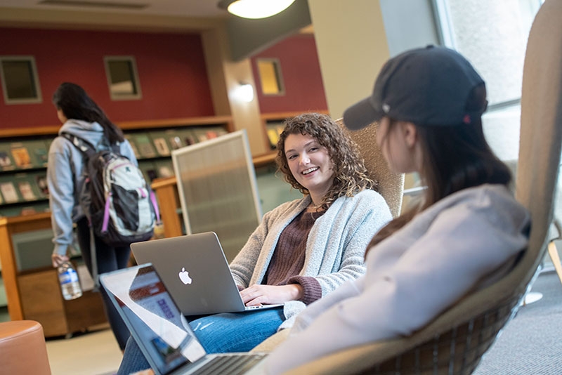 Two students sit in comfortable chairs in the periodical section of Schaffer Library, engaged in conversation