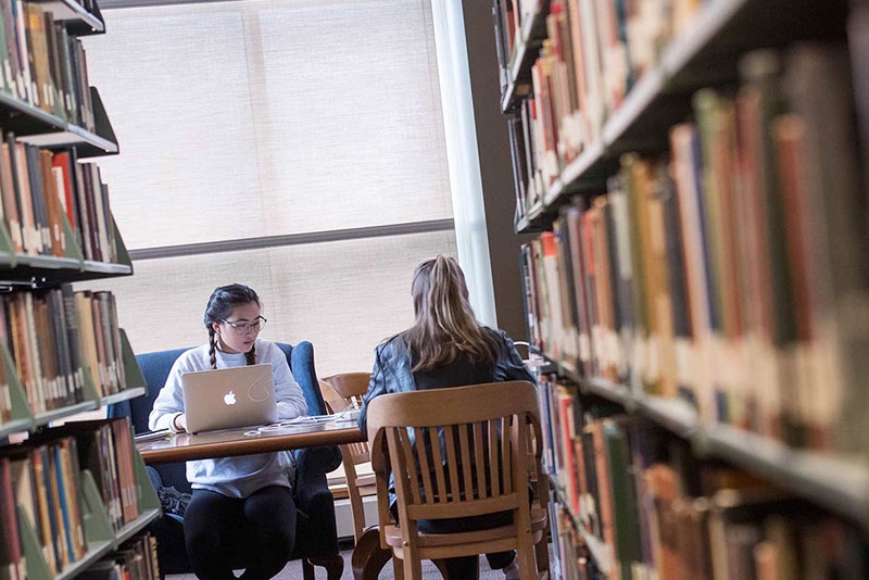 Two students sit at a wood desk in Schaffer Library, engaged in studying. To their left, a row of stacked books is visible.