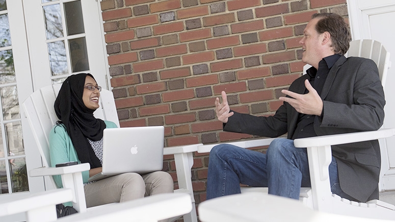 A student and professor converse while seated on the Lamont House front porch.