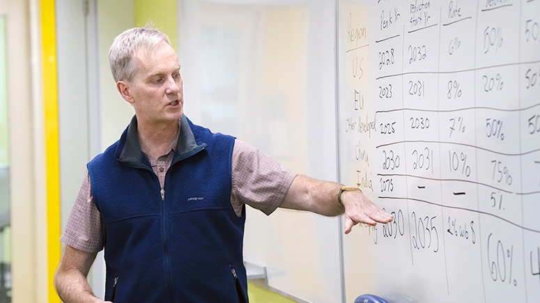 A professor points at a whiteboard displaying statistics on emissions released by various countries.