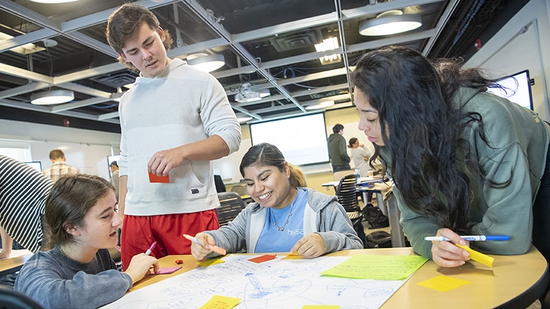 Students gather around a table in the Kelly Computing Lab classroom, studying a diagram spread out before them.